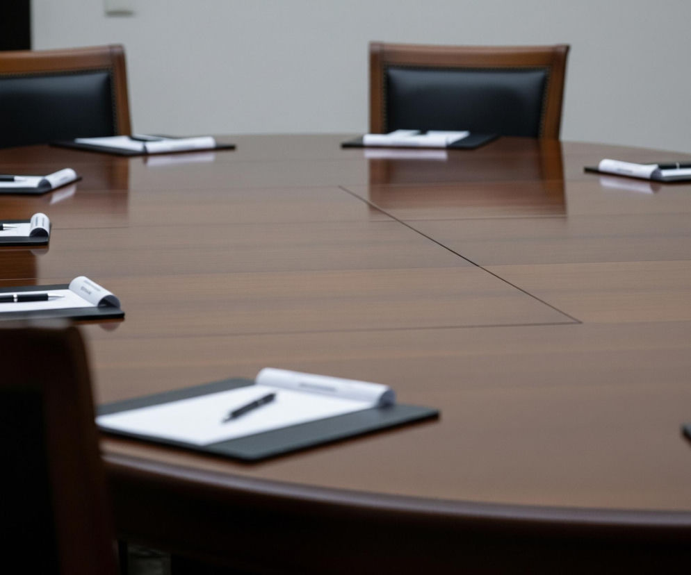 Długi, owalny stół konferencyjny z ciemnobrązowego drewna dominuje na zdjęciu. Wokół stołu ustawione są skórzane krzesła z brązowymi, drewnianymi oparciami i czarnymi siedziskami. Na stole, przed każdym krzesłem, leżą czarne podkładki z białymi kartkami i czarnymi długopisami. Na powierzchni stołu widoczne są subtelne linie, sugerujące jego składany charakter. Tło jest jasnoszare, w górnej części obrazu.