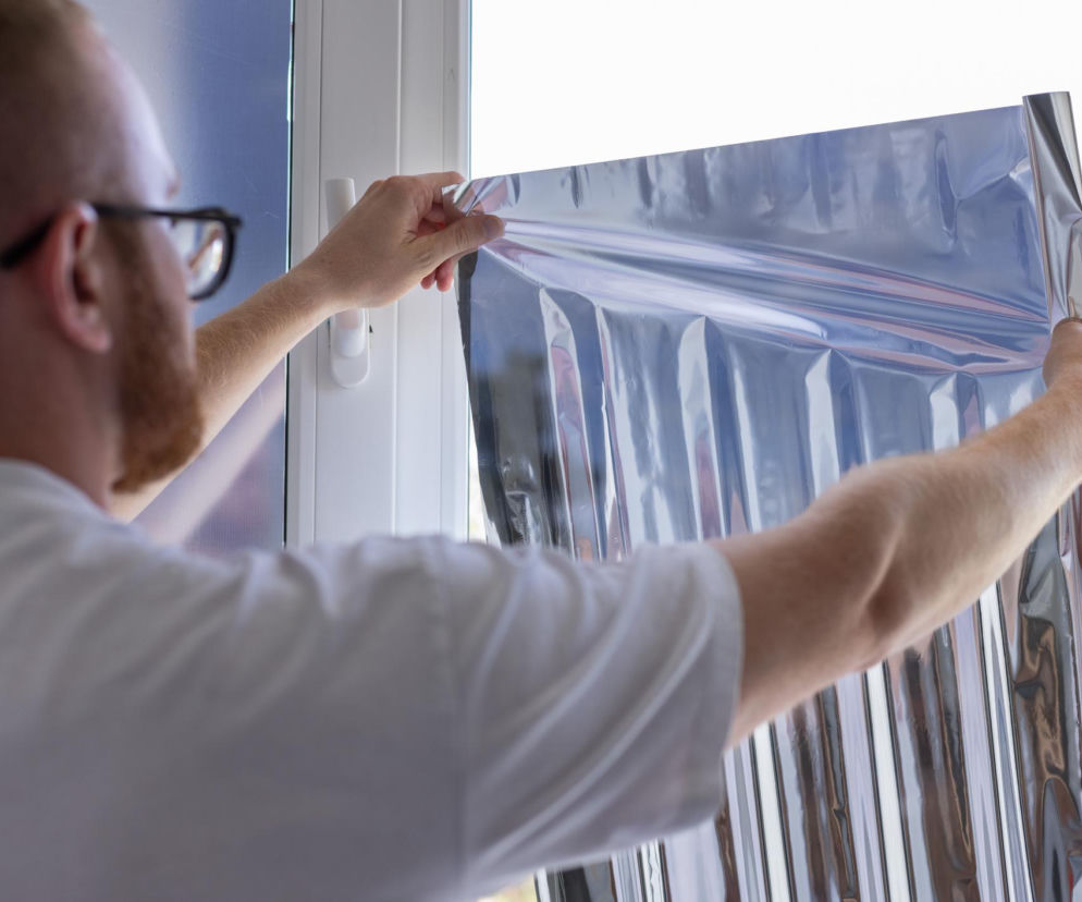 Mężczyzna w białej koszulce nakleja folię aluminiową na szybę okna. O trikach na upał przeczytasz na Muratordom.