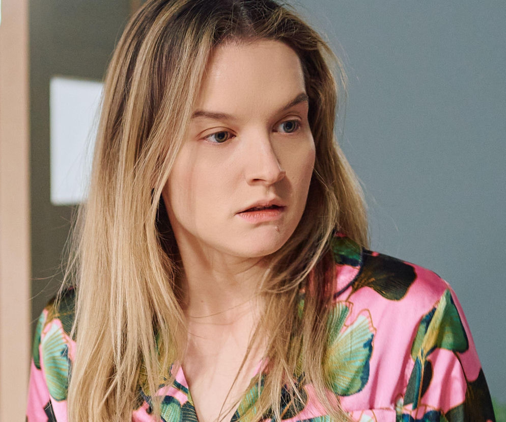 Aktorka Julia Chatys, wcielająca się w rolę Kasi w Na Wspólnej, wyraża na zdjęciu zaskoczenie lub oburzenie. Jej długie blond włosy rozpuszczone, ubrana jest w różową koszulę we wzory liściaste. Więcej o losach Kasi przeczytasz na portalu Super Seriale.