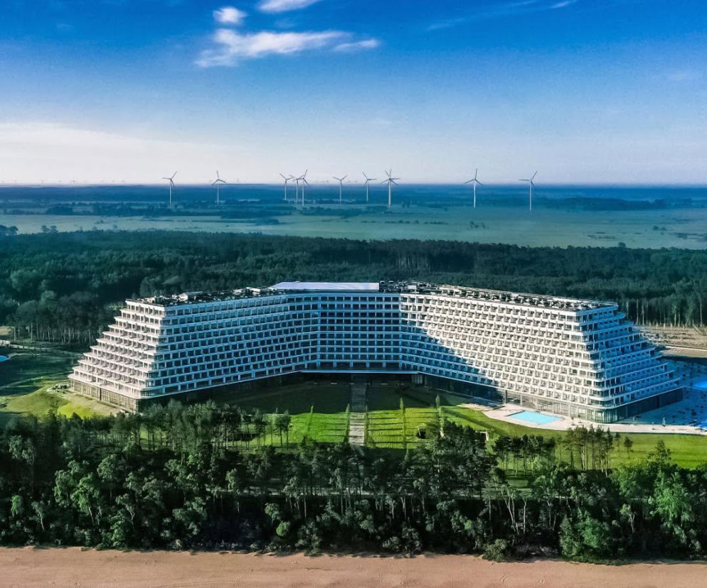 Ogromny, biały Hotel Gołębiewski w Pobierowie, widziany z lotu ptaka, otoczony zielonym lasem i piaszczystą plażą, z morzem w tle. W oddali widać turbiny wiatrowe. Więcej o tej gigantycznej inwestycji przeczytasz na Super Biznes.