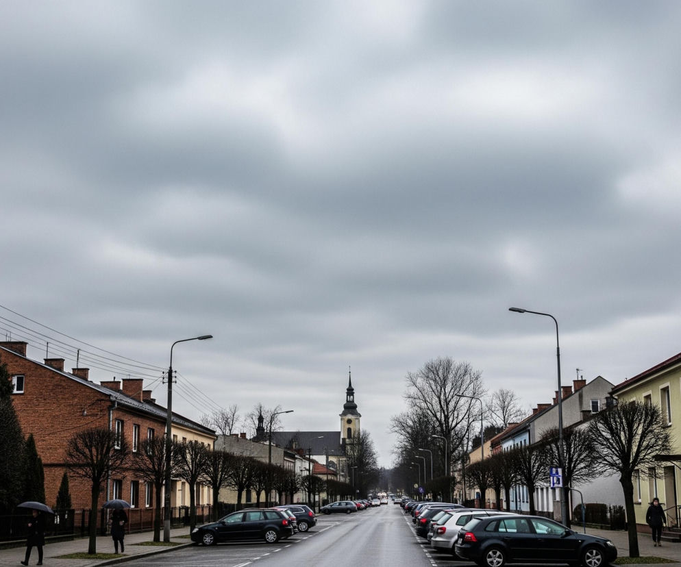 Długa, pusta ulica rozciąga się w głąb kadru, z ciemnym kościołem i jego strzelistą wieżą z zegarem widocznym na końcu perspektywy pod zachmurzonym, szarym niebem. Po obu stronach ulicy ciągną się dwupiętrowe budynki o różnorodnych elewacjach w odcieniach czerwieni, beżu i bieli, przed którymi rosną bezlistne drzewa i latarnie uliczne, z zaparkowanymi samochodami wzdłuż krawężników. Kilka postaci, w tym dwie z parasolami, przemieszcza się chodnikami, a kable telekomunikacyjne przecinają górną część lewej strony obrazu.