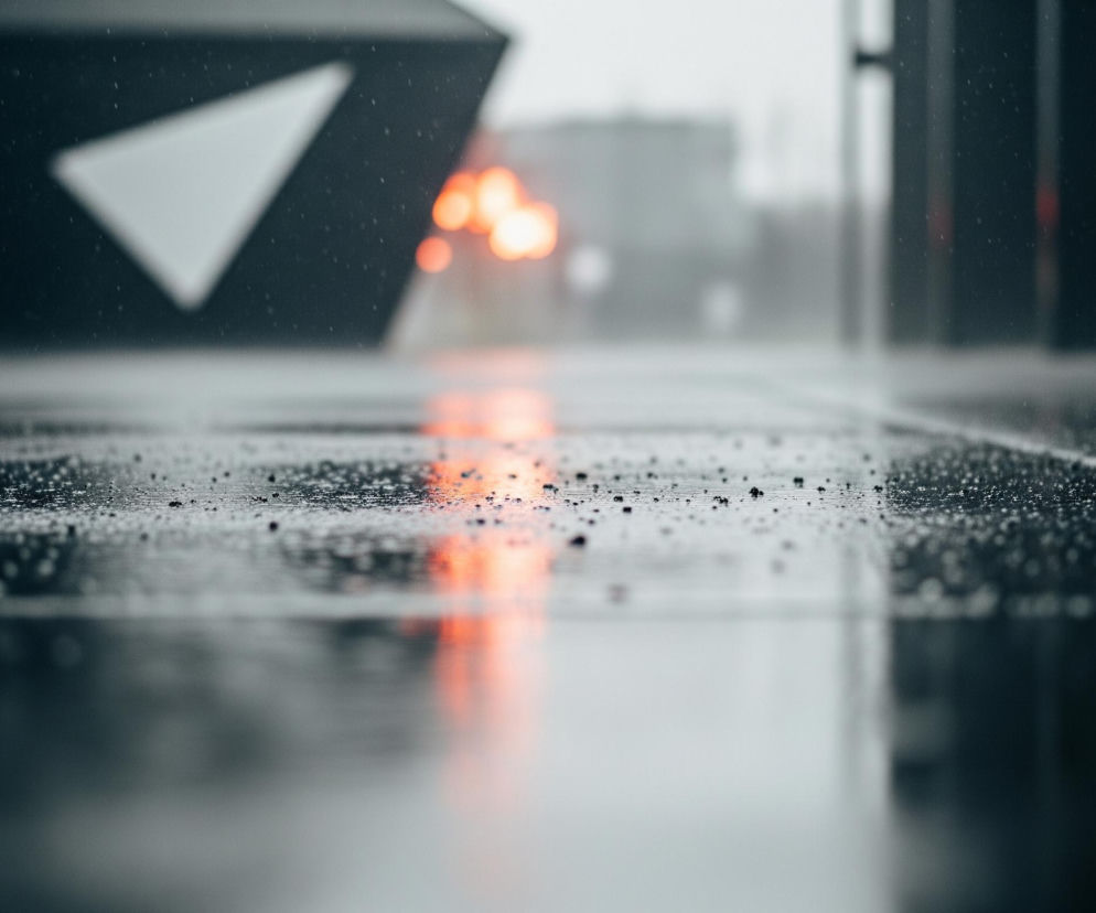 Uliczny chodnik lub jezdnia, pokryty deszczem i licznymi kroplami wody, rozciąga się w stronę horyzontu. W oddali, po lewej stronie, widoczna jest duża, ciemna struktura architektoniczna z białym, trójkątnym wzorem, odbijająca pomarańczowe światło. Po prawej stronie znajdują się rozmyte ciemne pionowe elementy, być może kolumny lub ramy, a tło wypełnia rozmyty, szarawy krajobraz miejski. Pomarańczowe światła, skupione pośrodku kadru, tworzą intensywne odblaski na mokrej powierzchni, dodając obrazowi ciepłego akcentu w dominującej szaro-niebieskiej palecie.