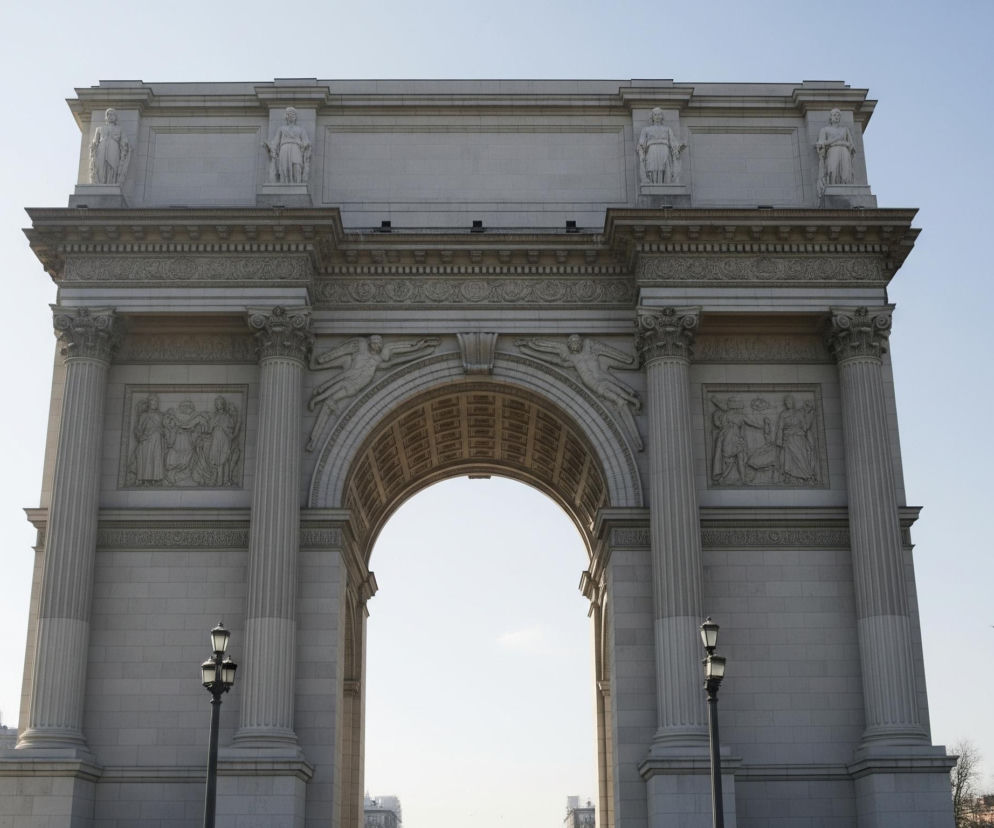 Duży, kamienny łuk triumfalny o jasnoszarym kolorze dominuje w centrum kadru, sięgając niemal od lewej do prawej krawędzi. Jego struktura jest bogato zdobiona rzeźbami, kolumnami z korynckimi kapitelami oraz detalami architektonicznymi, takimi jak płaskorzeźby przedstawiające postaci ludzkie i skrzydlate figury. W górnej części łuku, na płaskiej powierzchni, widoczne są cztery stojące posągi, po dwa po każdej stronie. Przejście pod łukiem odsłania jasne, bezchmurne niebo oraz niewielką część odległych budynków i drzew w dolnej, prawej części obrazu. Przed łukiem, po obu stronach, stoją czarne latarnie uliczne z trzema kloszami każda.