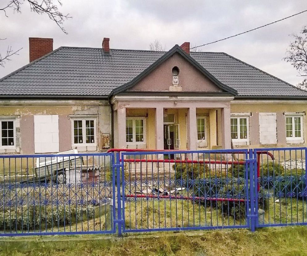 Stara wiejska szkoła przed gruntownym remontem, z łuszczącym się tynkiem, nowym dachem i wymienionymi oknami, za ogrodzeniem. Budynek czeka na przekształcenie w elegancką rezydencję, o czym więcej przeczytasz na naszym portalu.