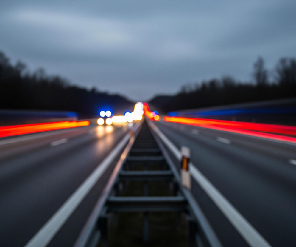 Na zdjęciu przedstawiona jest autostrada z rozmytymi światłami pojazdów. Dwie jezdnie rozdzielone są metalową barierką energochłonną, biegnącą przez środek kadru, od dołu ku górze. Światła tylne jadących samochodów tworzą czerwone smugi po lewej stronie drogi, natomiast światła przednie nadjeżdżających pojazdów widoczne są jako rozmyte, białe i niebieskie plamy w oddali, pośrodku kadru, tworząc także czerwoną smugę po prawej stronie. Otoczenie drogi stanowią ciemne, rozmyte drzewa po obu stronach, a niebo jest zachmurzone i szare.