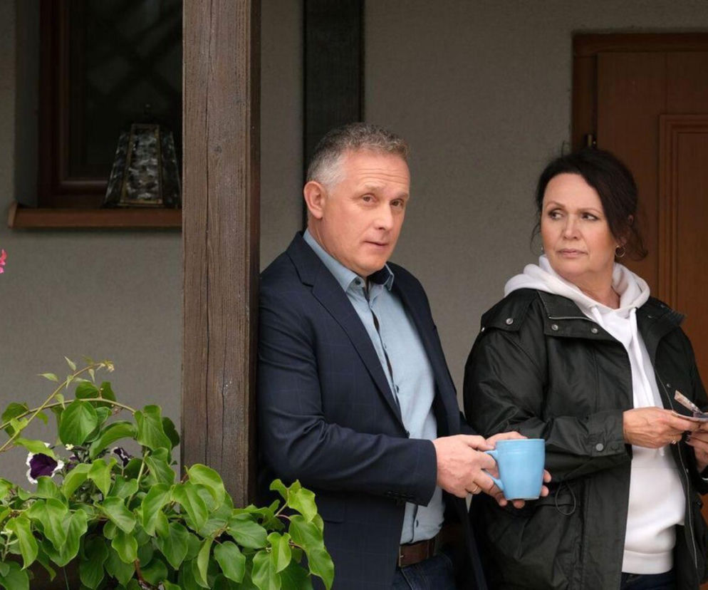 Artur Rogowski i Marysia Rogowska z M jak miłość stoją na ganku domu. Artur opiera się o drewniany słupek, trzymając niebieski kubek, patrzy prosto w kamerę. Marysia stoi obok, zamyślona, odwrócona lekko w prawo. Scena oddaje trudną rozmowę małżeńską, o której przeczytasz na Super Seriale.