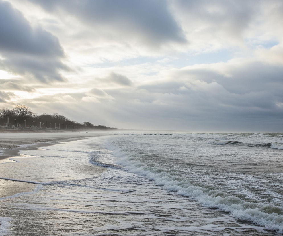 Szeroką, nadmorską plażę z wzburzonym morzem i pochmurnym niebem. Duże fale z białą pianą przybrzeżną rozbijają się na piasku w prawym dolnym rogu, tworząc wyraźne linie i mokre ślady na ciemnym, wilgotnym piasku. W oddali, wzdłuż linii brzegowej, widać aleję obsadzoną rzędami bezlistnych drzew i oświetloną latarniami, a dalej na horyzoncie z morza wystaje długie, ciemne molo lub falochron. Niebo wypełniają gęste, szare chmury, miejscami rozjaśnione promieniami słońca przebijającymi się przez zachmurzenie, tworząc jasne smugi światła.