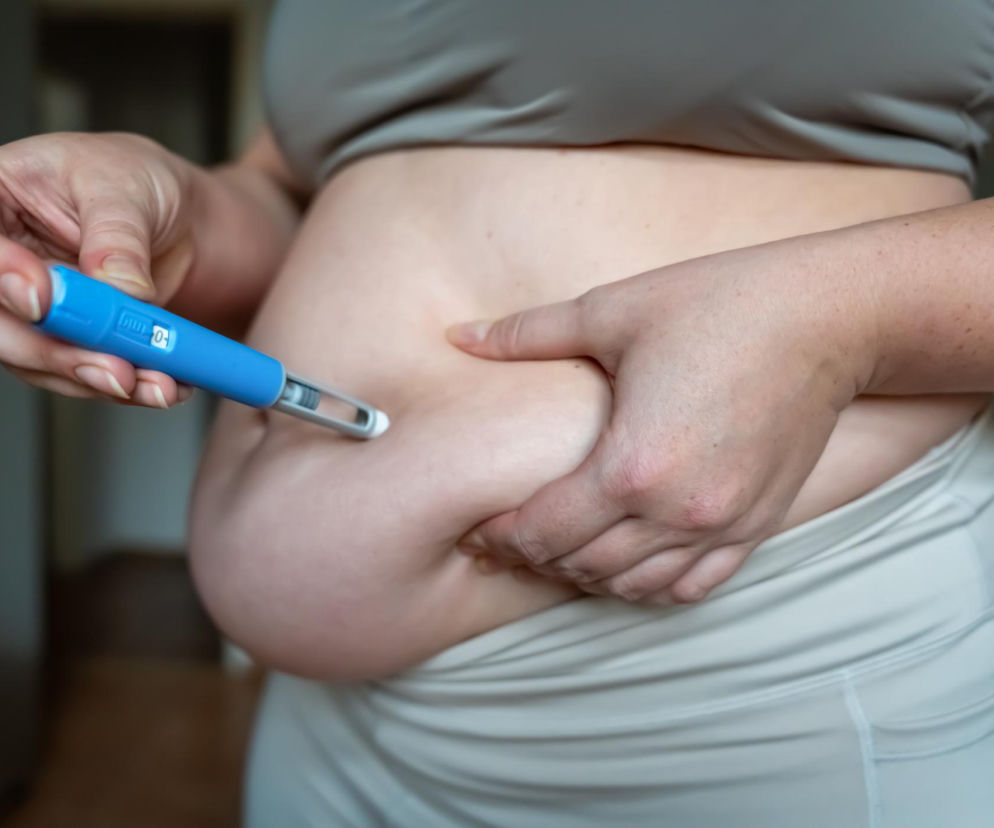 Osoba z nadwagą w jasnym biustonoszu i legginsach, wstrzykująca sobie lek odchudzający w fałd skóry na brzuchu za pomocą niebieskiego pena insulinowego. Na Poradnik Zdrowie znajdziesz informacje o nowych zastosowaniach leków GLP-1 po zawale serca.