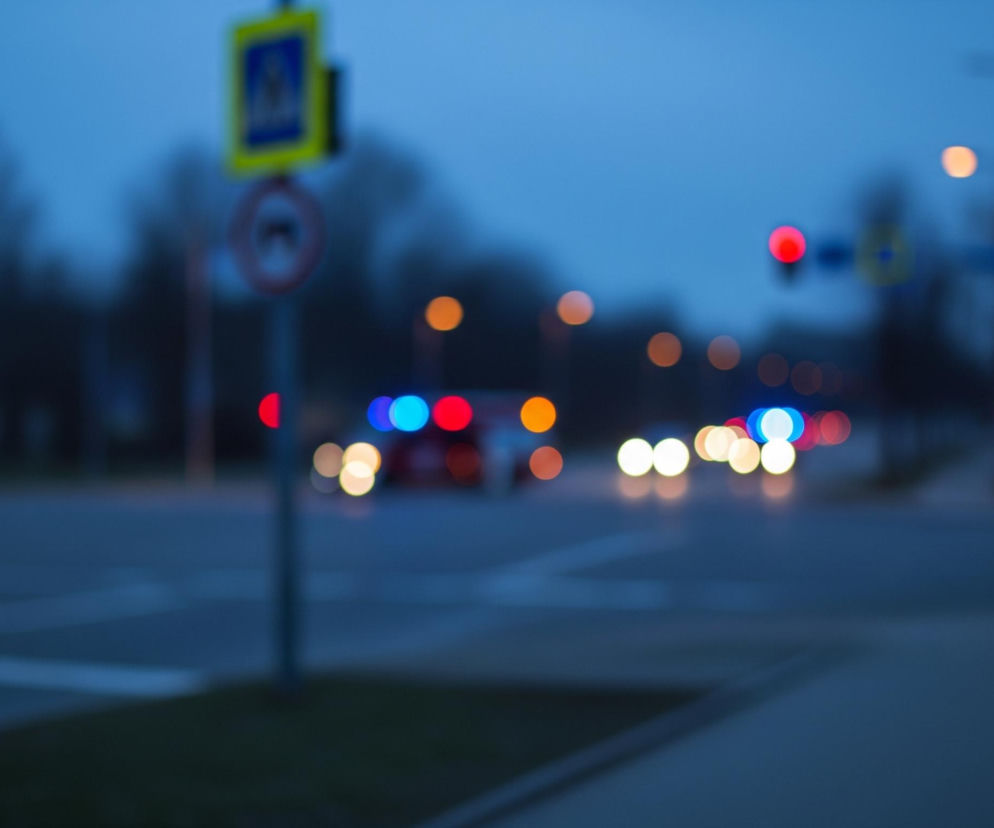 Rozmyty obraz przedstawia nocną scenę miejską, z głównym akcentem na kolorowe światła samochodów i uliczne lampy. Na pierwszym planie, po lewej stronie, widoczny jest słupek z dwoma znakami drogowymi: u góry żółty kwadratowy znak z symbolem pieszego, a poniżej okrągły znak zakazu skrętu w prawo. Dalej na horyzoncie, również po lewej stronie, zarysowane są drzewa ogołocone z liści. Ulica, częściowo widoczna na dole kadru, ma szarą nawierzchnię z białymi pasami. W centrum i po prawej stronie obrazu, liczne punkty świetlne o kolorach czerwonym, pomarańczowym, białym i niebieskim tworzą efekt bokeh, sugerując obecność pojazdów i sygnalizacji świetlnej. Dominujące kolory to różne odcienie niebieskiego i granatu, charakterystyczne dla zmierzchu lub nocy.