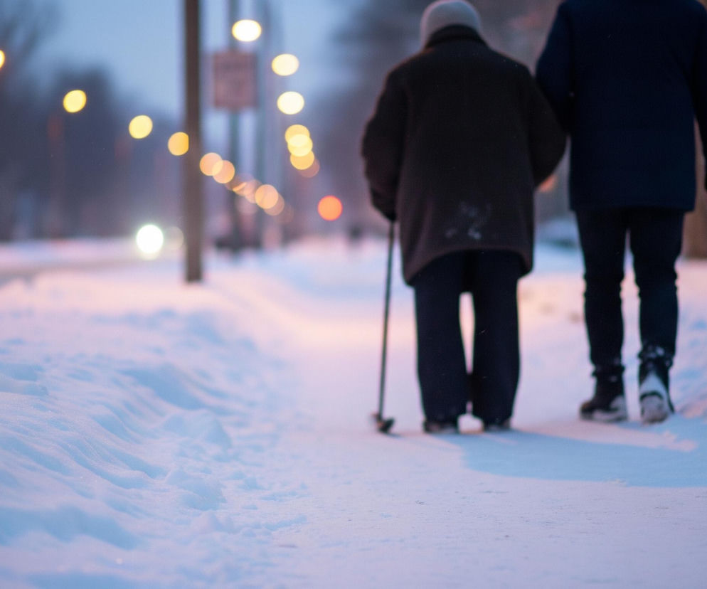 Dwie osoby idą po pokrytym śniegiem chodniku, oglądane od tyłu. Osoba po lewej, ubrana w ciemną kurtkę i jasną czapkę, opiera się o laskę, a obok niej idzie druga osoba w ciemnym płaszczu, wspierając ją. Tło jest rozmyte, z widocznymi światłami ulicznymi o ciepłym, żółtym odcieniu, rozciągającymi się w głąb obrazu. Śnieg na pierwszym planie jest delikatnie różowy i niebieski, co sugeruje wczesny wieczór lub poranek.