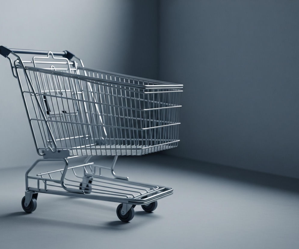 Metalowy wózek na zakupy, wykonany z cienkich, chromowanych prętów, stoi na gładkiej, jasnoszarej powierzchni, z prawym, dalszym rogiem lekko podniesionym. Jego koszyk jest pusty, a rączka pokryta jest czarnym, gumowym materiałem. Tło stanowi narożnik dwóch ścian w kolorze jasnoszarym, z widocznym cieniem w prawym górnym rogu. Całe ujęcie jest monochromatyczne, w odcieniach szarości i błękitu, z mocnym światłem padającym z lewej strony, tworzącym łagodny cień pod wózkiem.