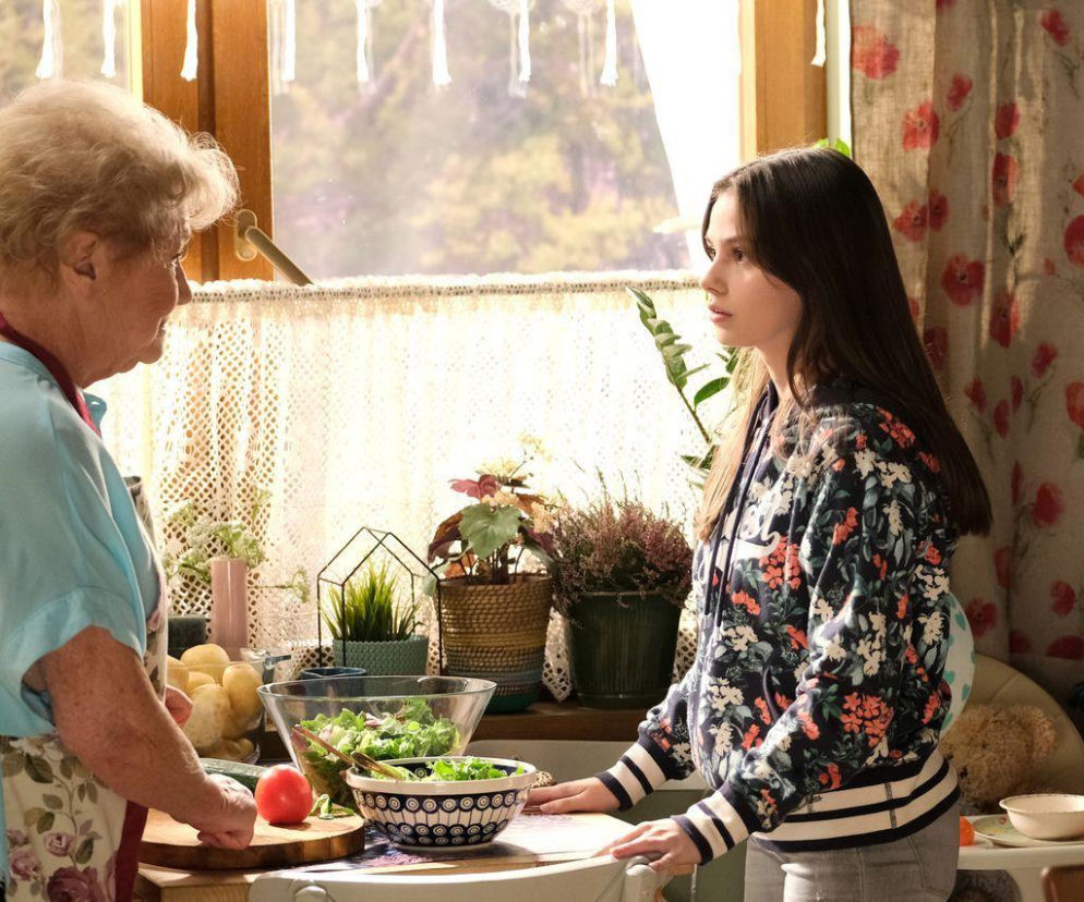 Basia Rogowska (Karina Woźniak) rozmawia z Barbarą Mostowiak (Teresa Lipowska) w kuchni, przy stole z warzywami i sałatkami. Młoda kobieta w kwiecistej bluzie z kapturem słucha starszej kobiety. Scena z serialu M jak miłość, więcej o niej na Super Seriale.