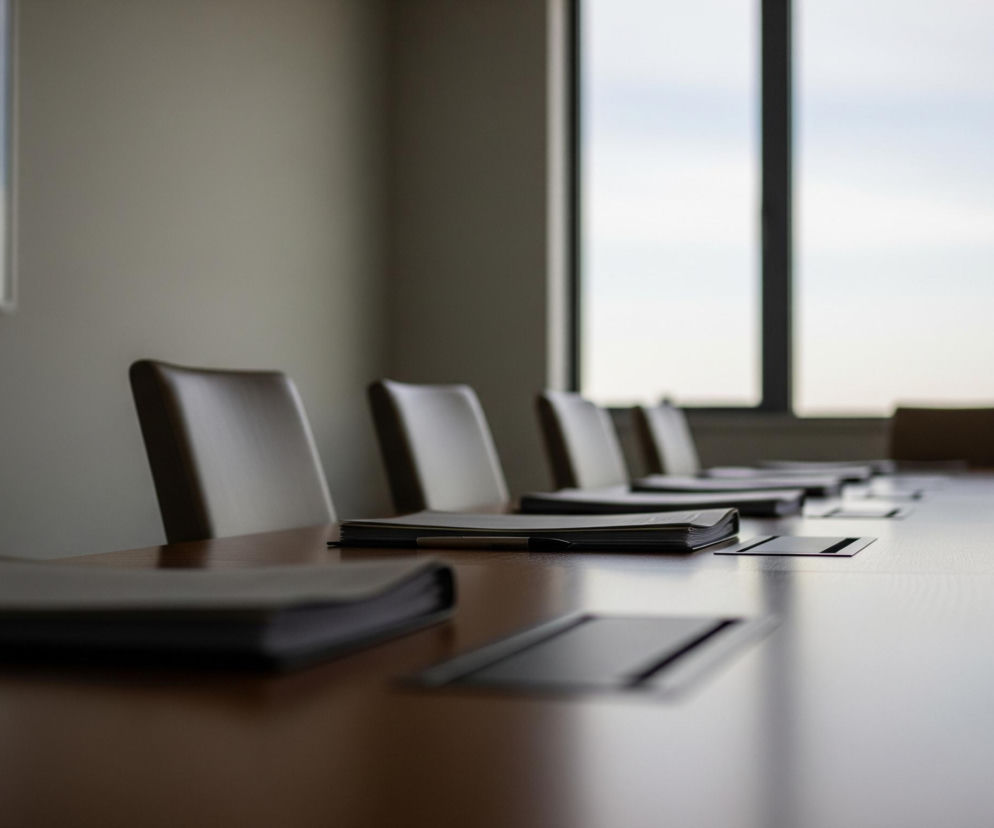 Długi, drewniany stół konferencyjny rozciąga się przez środek pomieszczenia, widoczny pod lekkim kątem od lewej dolnej krawędzi obrazu. Na stole ułożone są stosy czarnych lub ciemnoszarych dokumentów lub segregatorów, po jednym przed każdym z rzędów jasnych, skórzanych krzeseł, ustawionych po obu stronach stołu, z niewyraźnym tłem. Wzdłuż prawej ściany, w tle, widoczne jest duże okno z pionowymi szprosami, przez które wpada naturalne światło, rozjaśniając pomieszczenie i ukazując częściowo błękitne niebo. Na lewej ścianie, w tle, wisi prostokątny obraz w jasnej ramie, z niewyraźnym widokiem, prawdopodobnie zza okna.