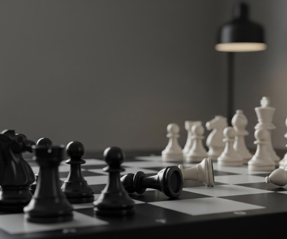 Na pierwszym planie, centralnie, widnieje szachownica z czarno-białymi figurami. Po lewej stronie, wzdłuż krawędzi szachownicy, ustawione są rzędy czarnych figur szachowych, a po prawej stronie widać białe figury. Dwie figury, jeden czarny pionek i jeden biały goniec, leżą przewrócone na szachownicy, bliżej środka kadru. W tle po lewej stronie znajduje się jasne okno, a po prawej stronie, w oddali, widać czarną lampę o rozproszonym świetle.