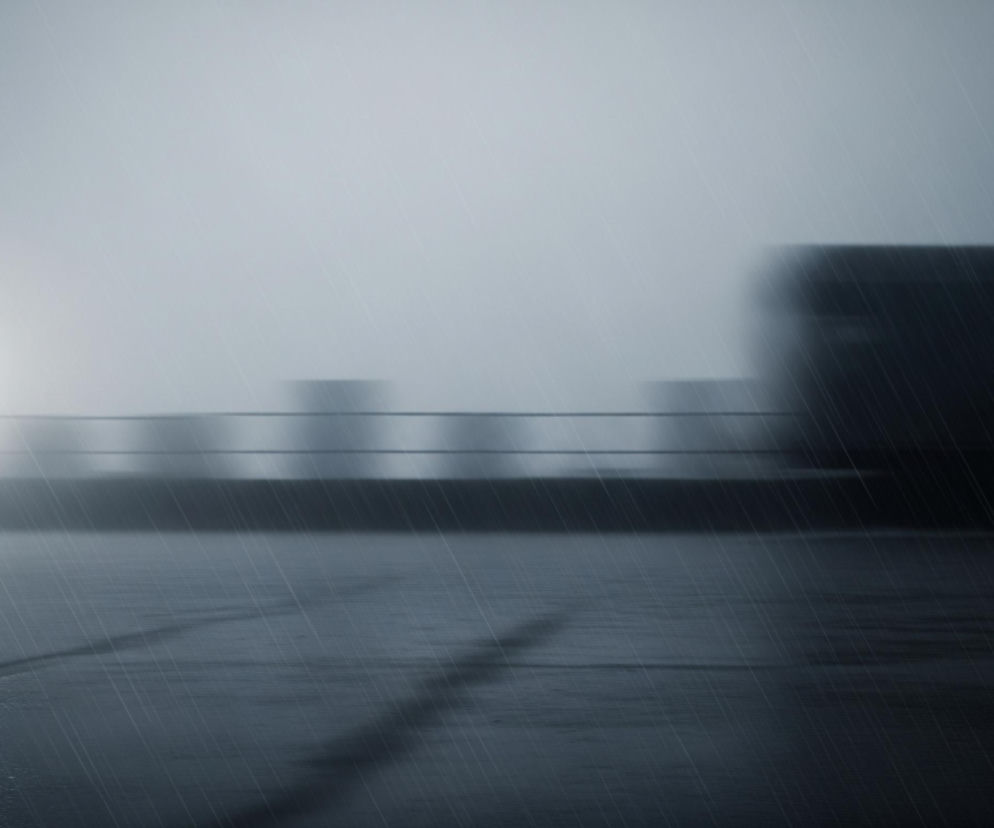 Ciemną, deszczową scenerię, gdzie wyraźnie widać padające ukośnie krople deszczu. Na pierwszym planie znajduje się mokra, odbijająca światło powierzchnia, na której pojawia się smugowate odbicie jasnego światła z lewej strony. W tle, na horyzoncie, rozciąga się niewyraźny zarys miejskiego krajobrazu, z kilkoma rozmazanymi budynkami i długim, ciemnym kształtem przypominającym ciężarówkę po prawej stronie. Całość utrzymana jest w chłodnych odcieniach szarości i błękitu, z dominującą atmosferą pochmurnego dnia lub zmierzchu.