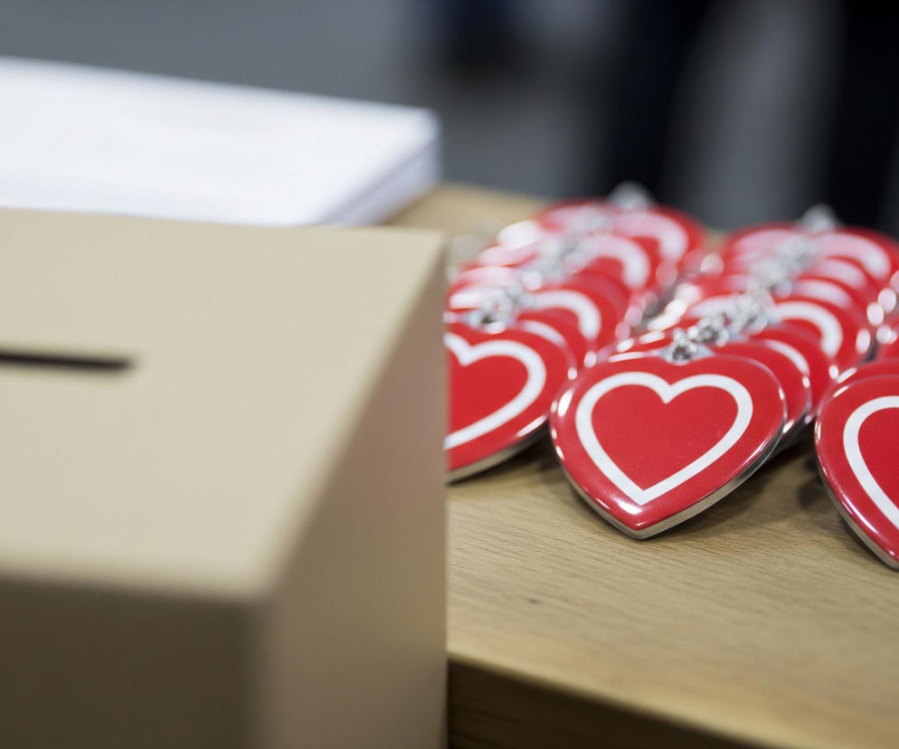 Na pierwszym planie, z lewej strony, widoczne jest beżowe pudełko z poziomym, ciemnym otworem na górze, częściowo zamazane. Po prawej stronie pudełka, na brązowym, drewnianym blacie, ułożone są w rzędach liczne czerwone breloczki w kształcie serc, każdy z białą obwódką wewnątrz, przymocowane do srebrnych łańcuszków kulkowych. Tło jest rozmyte, ukazując jaśniejsze i ciemniejsze obszary, sugerujące obecność innych obiektów lub przestrzeni.