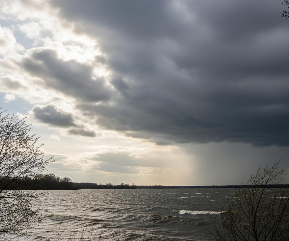 Kadr przedstawia wzburzoną taflę jeziora pod zachmurzonym niebem. Na pierwszym planie, po lewej stronie, widoczne są gałęzie drzewa bez liści, a po prawej delikatne gałązki z młodymi pąkami. Lewa strona nieba jest częściowo jasna, z przebijającymi się promieniami słońca, podczas gdy prawa strona jest pokryta ciemnymi, burzowymi chmurami, z widocznym opadem deszczu w oddali. Horyzont tworzy ciemny pas zalesionego brzegu, a woda jest ciemnoszara, z licznymi falami i białymi grzywami, świadczącymi o silnym wietrze.