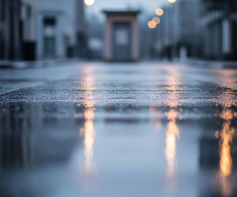 Mokra ulica, pokryta drobnymi kroplami wody, odbija rozmyte światła w odcieniach pomarańczu i błękitu, tworząc smugi wzdłuż perspektywy. W tle, niewyraźne zarysy budynków stoją po obu stronach drogi, a pojedyncza, beżowa budka telefoniczna znajduje się na środku, delikatnie zarysowana. Uliczne lampy, widoczne jako okrągłe, rozmyte plamy światła, zawisają nad całością, rzucając swe blade refleksy na powierzchnię.