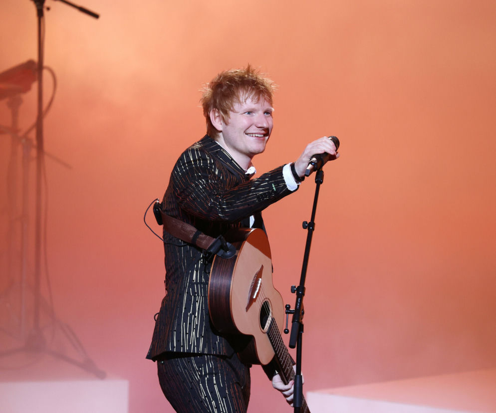 Uśmiechnięty Ed Sheeran z gitarą na scenie podczas koncertu. O kontrowersjach wokół muzyka przeczytasz w serwisie Eska.