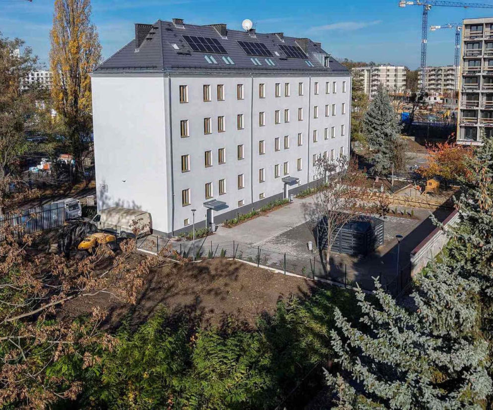 Nowoczesna kamienica z jasną elewacją i panelami fotowoltaicznymi na dachu, otoczona zielenią i nowymi nasadzeniami na Przedmieściu Oławskim we Wrocławiu. Zdjęcie ilustruje inwestycje zgodne z Zielonym Standardem Budynku, o którym można przeczytać na portalu Architektura Murator Plus.