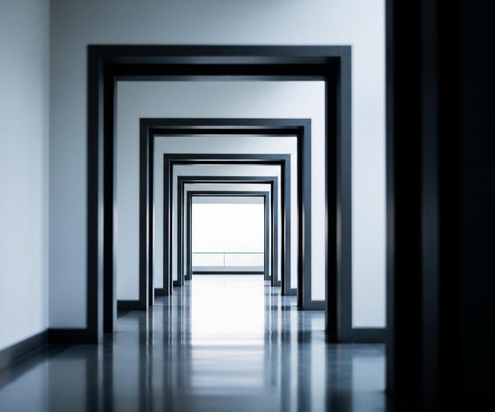 W długim, symetrycznym korytarzu widać szereg prostokątnych, ciemnych ościeżnic, które stają się coraz mniejsze w oddali, tworząc efekt tunelu. Ściany są jasnoszare, a podłoga, wykonana z błyszczącego, ciemnego materiału, odbija światło i zarysy ościeżnic. Na końcu korytarza, przez ostatnie, najmniejsze przejście, widać intensywnie jasne światło, co sugeruje otwartą przestrzeń lub okno.