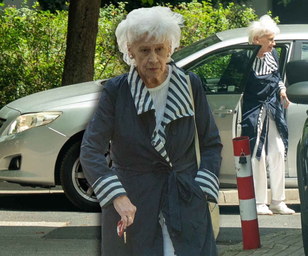 Krystyna Loska w granatowym płaszczu i białych spodniach. Obok miniatura gwiazdy przy aucie. O stylu ikony dowiesz się w SE.