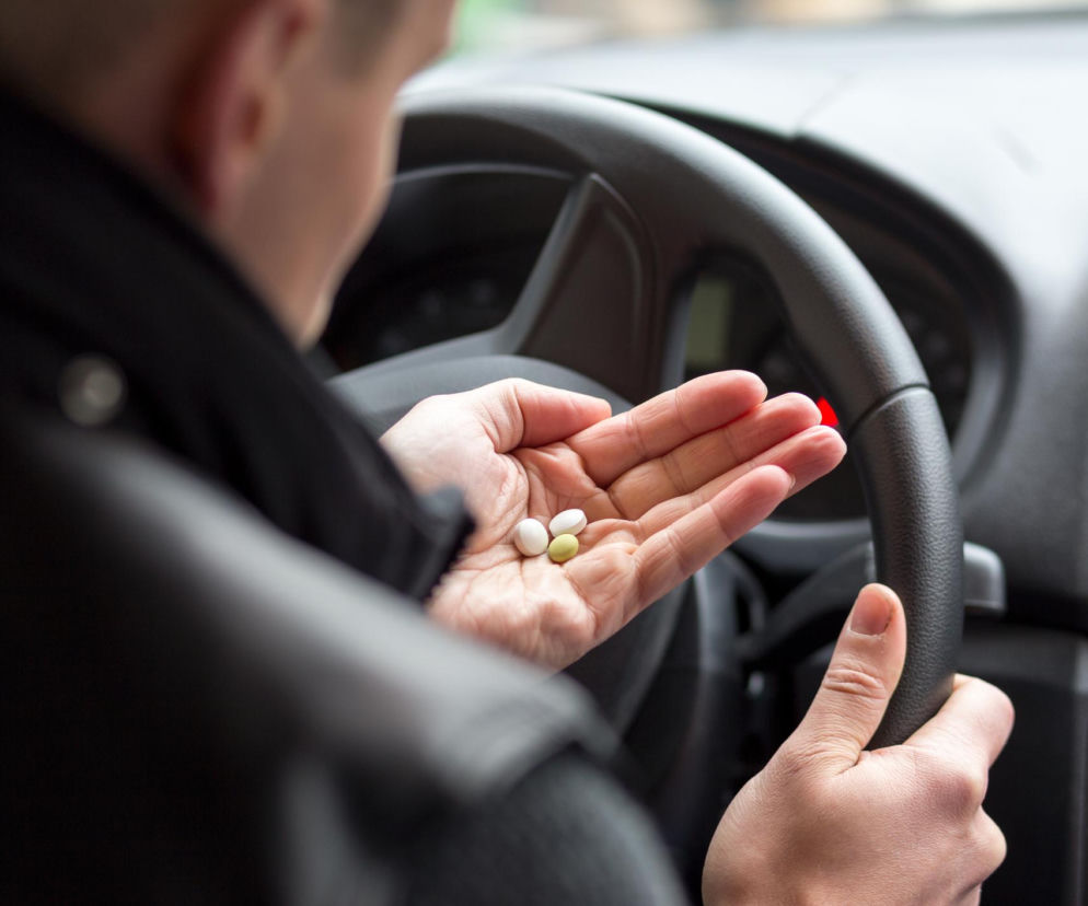 Mężczyzna za kierownicą samochodu trzyma w otwartej dłoni trzy tabletki: dwie białe i jedną żółtą. Zdjęcie ilustruje temat wpływu leków na zdolność prowadzenia pojazdów. O tym, jak leki wpływają na prowadzenie auta, przeczytasz więcej na Poradnik Zdrowie.