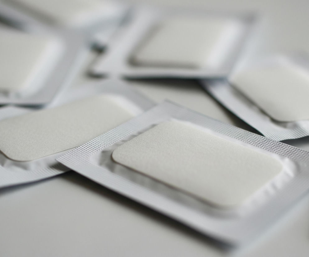 Sześć pojedynczo zapakowanych, prostokątnych produktów leży rozłożonych na jasnoszarej powierzchni. Każdy produkt składa się z białego, lekko wypukłego elementu, osadzonego w srebrnym opakowaniu z widocznymi zgrzewami w drobną kratkę na krawędziach. Obiekty są rozłożone nieregularnie, a niektóre częściowo zachodzą na siebie, z centralnym elementem w wyraźnym fokusie i pozostałymi delikatnie rozmytymi.