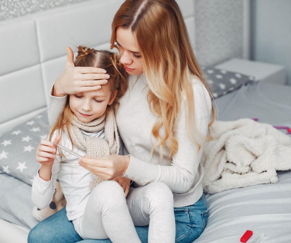 Mama sprawdza czoło chorej córki trzymającej termometr. O objawach grypy u dzieci przeczytasz na SE.
