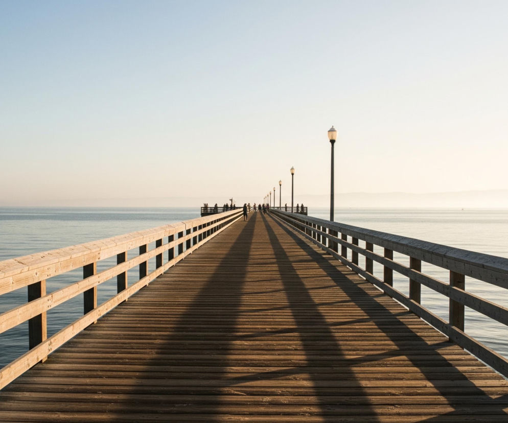 Długie, drewniane molo rozciąga się w głąb spokojnego morza, tworząc wyraźną perspektywę prowadzącą do horyzontu. Po obu stronach mola widoczne są drewniane balustrady, a na jego powierzchni padają długie cienie od latarni rozmieszczonych wzdłuż konstrukcji. W oddali, na końcu mola, gromadzi się grupa ludzi, a na powierzchni wody unosi się kilka małych łodzi. Niebo jest jasne, o delikatnym przejściu kolorów od jasnoniebieskiego u góry do niemal białego nad horyzontem, co sugeruje wschód lub zachód słońca.
