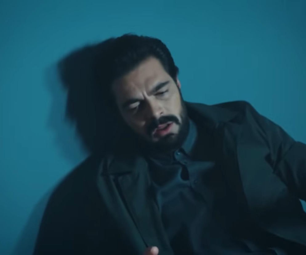 Yaman Kirimli z brodą i wąsami, w ciemnej koszuli i marynarce, osunął się na niebieską ścianę, jego twarz wyraża cierpienie i dezorientację. Scena z serialu Dziedzictwo, gdzie Yaman został otruty. Więcej o losach bohatera przeczytasz na portalu Super Seriale.