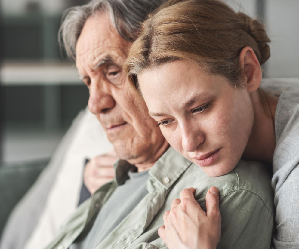 Zasmucona młoda kobieta obejmuje starszego, pogrążonego w myślach mężczyznę, symbolizując wsparcie w chorobie. Obrazek ilustruje tematykę nowotworów i choroby Alzheimera, o której przeczytasz na Poradnik Zdrowie.