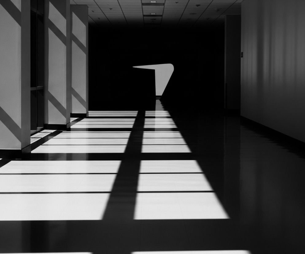 Długi korytarz ukazuje intensywny kontrast światła i cienia, tworząc dramatyczną kompozycję w odcieniach czerni i bieli. Z lewej strony światło wpada przez pionowe szczeliny, rzucając na podłogę prostokątne pasy jasnych refleksów, które zbiegają się ku odległemu, tajemniczemu zakończeniu korytarza. Głębia obrazu pogłębia się stopniowo w ciemność, a na horyzoncie widoczny jest abstrakcyjny, biały kształt, przypominający zakrzywiony znak zapytania lub fragment oświetlonej ściany, który przełamuje jednolitą czerń. Prawa strona korytarza pozostaje w głębokim cieniu, z jedynie subtelnymi przejściami tonalnymi na ścianie, podkreślającymi perspektywę i prowadzącymi wzrok w głąb pomieszczenia.