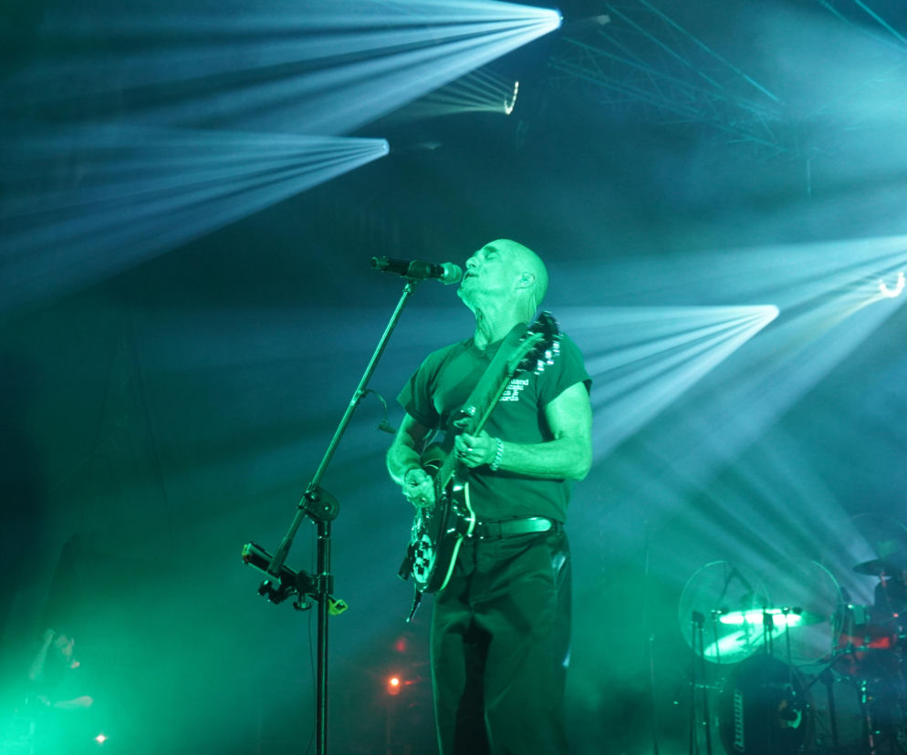 Artur Rojek, były wokalista zespołu Myslovitz, na scenie podczas koncertu, śpiewający do mikrofonu i grający na gitarze elektrycznej, oświetlony zielono-niebieskim światłem reflektorów. Kontekst dotyczy jego wypowiedzi o trudnościach kariery i niekorzystnych kontraktach, o czym można przeczytać na naszym portalu.
