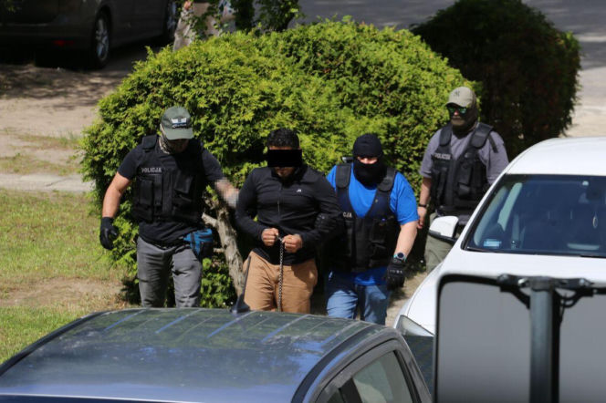Aresztowany Elnur A. w kajdankach i z zasłoniętą twarzą, prowadzony przez funkcjonariuszy policji w kamizelkach taktycznych, na tle zielonych krzewów i samochodów, w związku z zarzutem zabójstwa w Białej Podlaskiej. Więcej informacji o tymczasowym areszcie znajdziesz na naszym portalu.