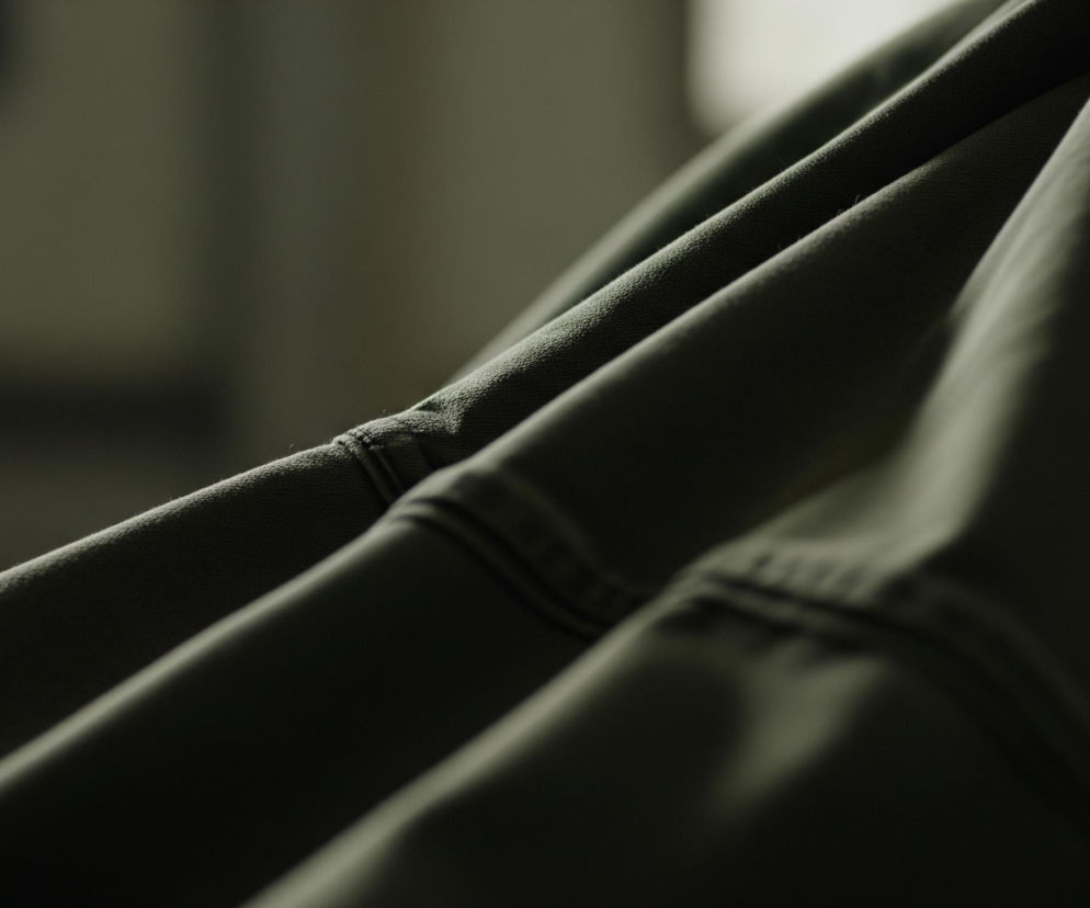 Fragment tkaniny w kolorze ciemnozielonym lub oliwkowym, z widocznymi zagnieceniami i fałdami, jest ułożony ukośnie, dominując w kadrze. Lewa górna część materiału charakteryzuje się bardziej wyraźną teksturą, z widocznymi szwami biegnącymi równolegle do fałd, podkreślającymi jego strukturę. Oświetlenie akcentuje te fałdy, tworząc miękkie cienie i rozjaśnienia, co nadaje tkaninie głębi i trójwymiarowości. Tło jest rozmyte, utrzymane w podobnej tonacji barwnej, co tkanina, choć jaśniejsze w górnej części.