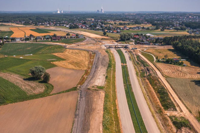Widok z góry na plac budowy drogi ekspresowej wśród pól i domów. O inwestycjach drogowych przeczytasz na Muratorplus.