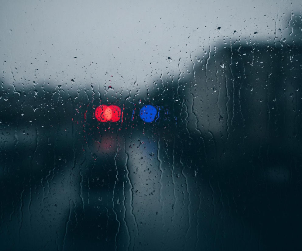 Szyba pokryta jest licznymi kroplami deszczu, które spływają w dół, tworząc smugi na jej powierzchni. Rozmyte tło przedstawia budynki po prawej stronie oraz drzewa i niebo po lewej i w górnej części, wszystkie w odcieniach szarości i ciemnego błękitu. Na wysokości horyzontu widać dwa jaskrawe, rozmazane światła: jedno czerwone po lewej i jedno niebieskie po prawej, kontrastujące z ogólnym, ciemnym i ponurym nastrojem obrazu.