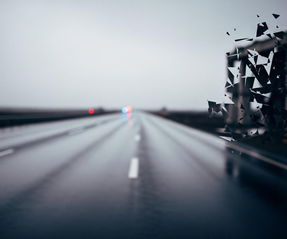Centralnie na zdjęciu znajduje się szeroka, mokra jezdnia autostradowa z białymi pasami ruchu, biegnąca w oddali pod lekkim kątem w lewo. Na horyzoncie widoczne są rozmyte światła samochodów, głównie czerwone i niebieskie, odbijające się od wilgotnej nawierzchni. Po prawej stronie, w tle, widnieje częściowo rozpadający się budynek o ciemnych, rozmytych konturach, z którego odrywają się liczne, ostre, czarne i szare fragmenty w kształcie trójkątów i wielokątów, rozrzucane w prawą stronę. Lewa strona zdjęcia przedstawia rozmyte pasy ruchu i bariery drogowe, zlewające się z szarym, zamglonym niebem.