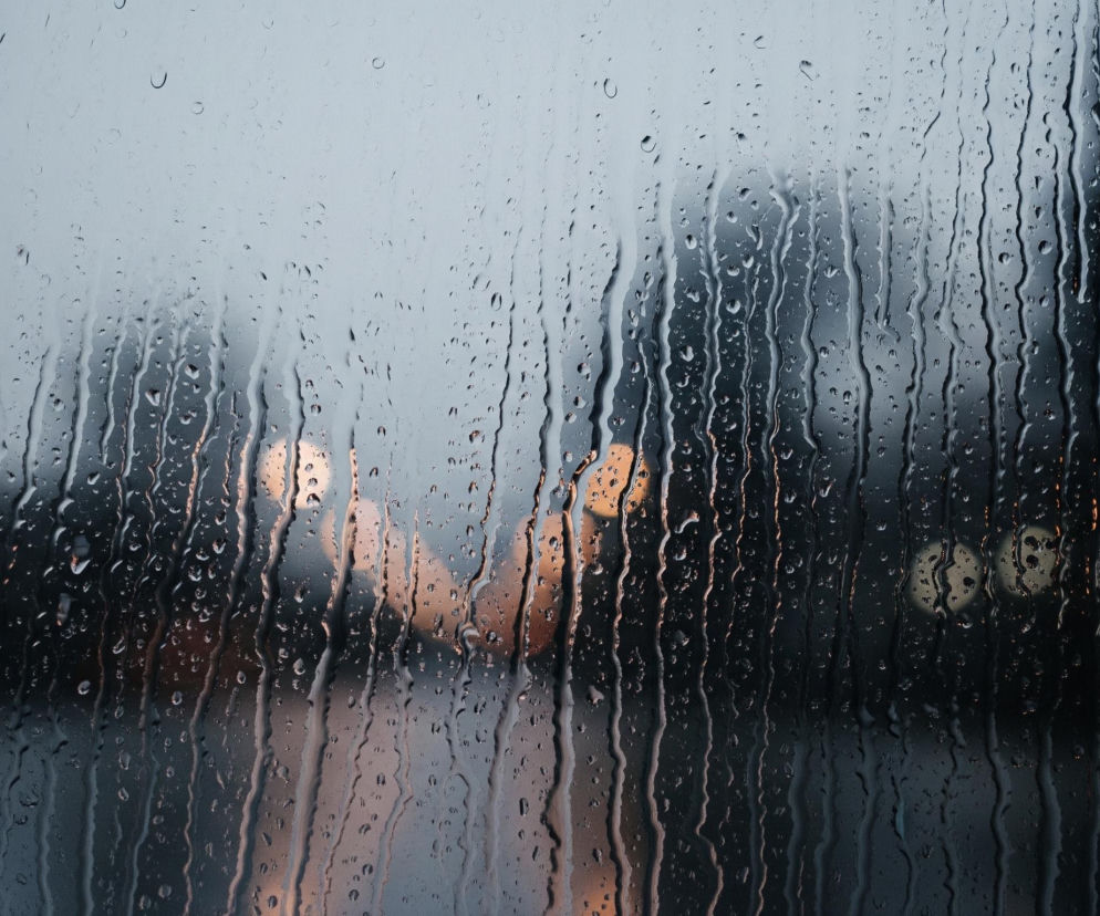Szybę pokrytą licznymi kroplami deszczu i smugami wody spływającymi pionowo. Za szybą widoczny jest rozmyty, miejski krajobraz, gdzie ciemne, pionowe kształty budynków zlewają się z szarym niebem w górnej części. W tle, na wysokości około połowy kadru, wyróżniają się jaskrawe, okrągłe plamy światła w odcieniach pomarańczowego i żółtego, będące prawdopodobnie latarniami ulicznymi lub światłami pojazdów. Całość ma chłodną, deszczową atmosferę.