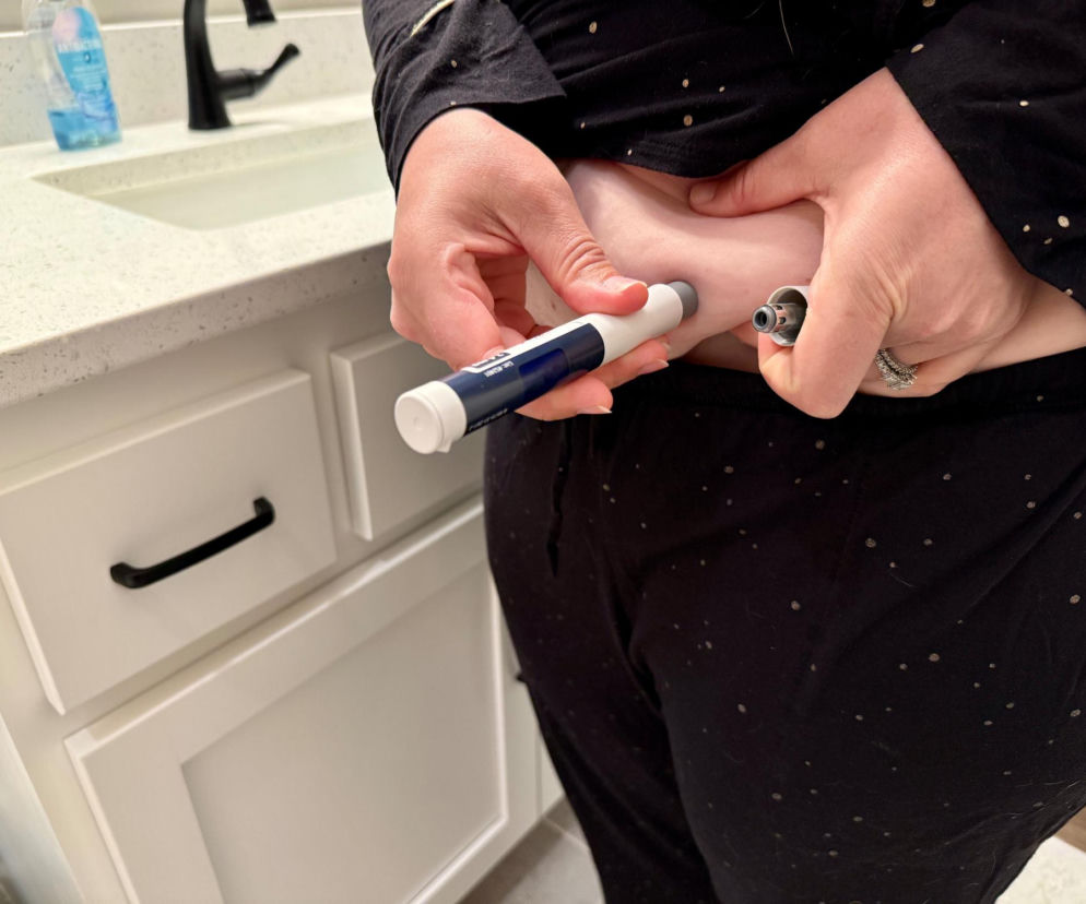 Osoba w ciemnej piżamie, w łazience, samodzielnie robiąca zastrzyk na odchudzanie z użyciem pena, chwytając fałd skóry brzucha. Na portalu Poradnik Zdrowie można przeczytać, czy suplementy są konieczne przy stosowaniu leków GLP-1.