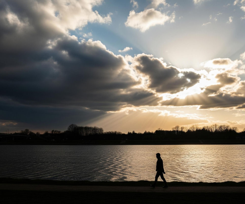 Ciemna sylwetka osoby idącej w lewo zajmuje centralną część dolnej połowy kadru, poruszając się wzdłuż ciemnego pasa ziemi, nad zbiornikiem wodnym o falującej powierzchni. Poziom wody rozjaśnia się w kierunku prawej strony obrazu, odzwierciedlając złote odcienie zachodzącego słońca, którego promienie przebijają się przez warstwy chmur. Niebo od lewej górnej części wypełniają ciemne, burzowe chmury, przechodzące w jaśniejsze i bardziej puszyste w kierunku prawej, skąd rozchodzą się jasne pasma światła. Na horyzoncie, po drugiej stronie wody, widać ciemne sylwetki drzew, a w oddali po lewej stronie majaczy fragment mostu.