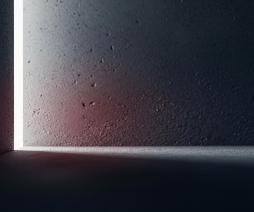 Ciemne, teksturowane pomieszczenie z jaśniejszym światłem emanującym z wąskiej, pionowej szczeliny po lewej stronie. Światło tworzy ostry cień wzdłuż dolnej części ściany i na podłodze. Powierzchnia ściany i podłogi jest szara, niejednolita, z widocznymi drobnymi nierównościami i rozproszonymi jasnymi plamkami, a także lekkim, czerwonawym odcieniem w dolnej części ściany, blisko źródła światła.