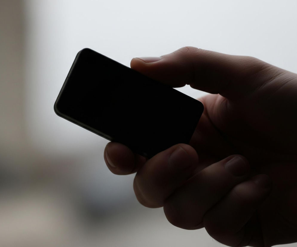 Centralnie w kadrze znajduje się ciemna dłoń trzymająca prostokątny, czarny przedmiot przypominający telefon komórkowy. Palce obejmują przedmiot, a jego ekran jest całkowicie ciemny, bez widocznego obrazu. Tło jest rozmyte, jasnoszare, z delikatnymi różnicami w odcieniach, sugerującymi naturalne światło padające z tyłu.