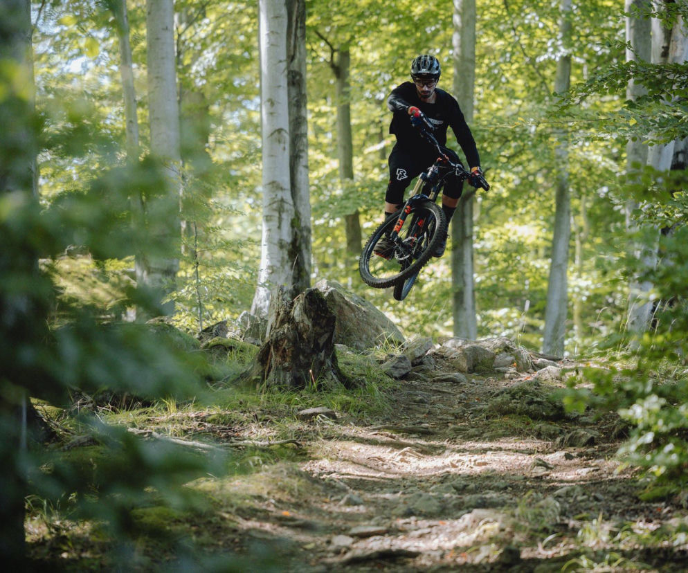 Rowerzysta w kasku skacze na rowerze enduro nad kamieniami w lesie. O zawodach w Szklarskiej Porębie czytaj na SE Superbiz.
