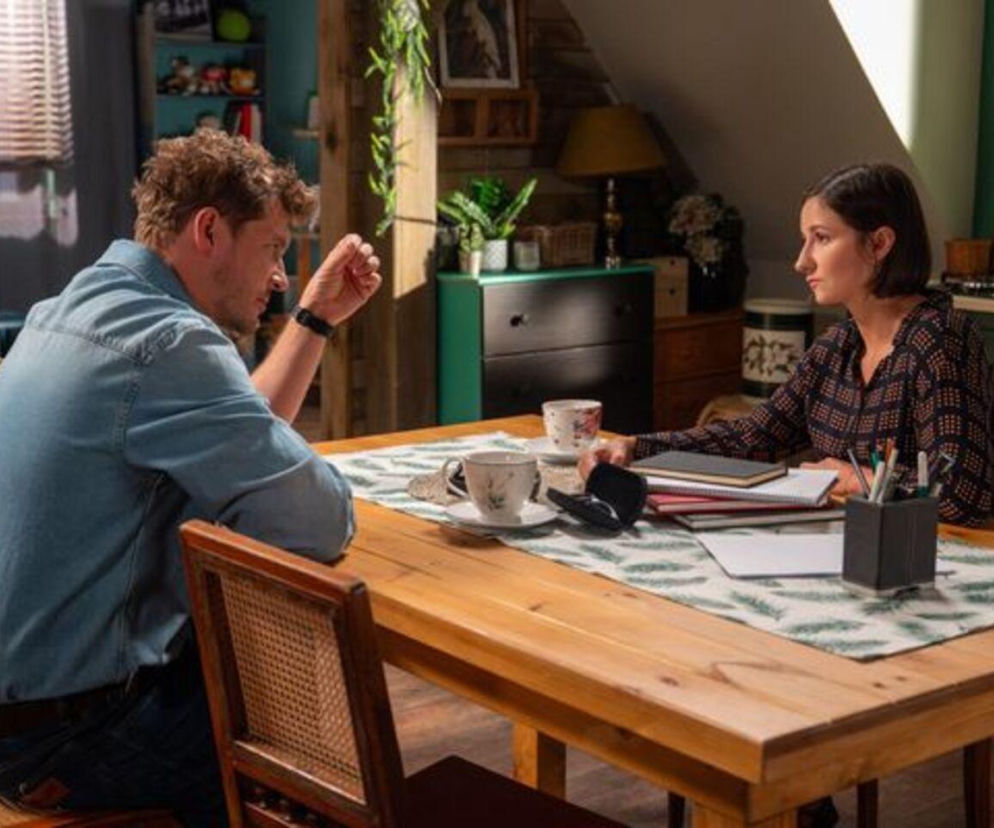 Mikołaj Roznerski jako Marcin Chodakowski i Amanda Mincewicz jako Agnes siedzą przy drewnianym stole w kuchni. Marcin w dżinsowej koszuli gestykuluje dłonią, a Agnes w ciemnej wzorzystej bluzce słucha go. Na stole leżą otwarte zeszyty i kubki, co zapowiada dramatyczne wydarzenia w M jak miłość. Więcej na temat serialu przeczytasz na portalu Super Seriale.