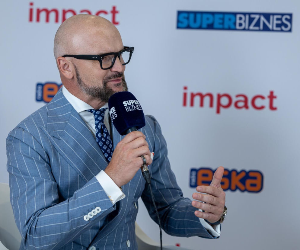 Rafał Brzoska, prezes InPostu, w niebieskim garniturze w prążki, okularach, podczas wywiadu. Trzyma mikrofon z logo Super Biznes, gestykulując. W tle widać logo Impact oraz Super Biznes. O opiniach Brzoski o polskiej gospodarce przeczytasz na Super Biznes.