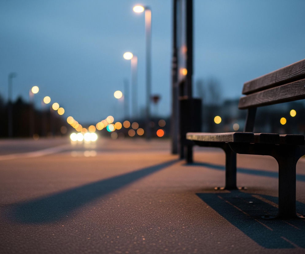 Na pierwszym planie, z prawej strony, stoi ciemnobrązowa ławka z drewnianymi deskami tworzącymi siedzisko i oparcie, rzucająca długi cień na szary chodnik. Przed ławką, po lewej stronie, rozciąga się asfaltowa ulica z dwoma białymi pasami ruchu, po której w oddali widać rozmyte, jasne światła samochodów. W tle, wzdłuż ulicy, widać szereg rozmazanych, żółtych lamp ulicznych, a niebo ma odcień ciemnego błękitu przechodzącego w szarość.