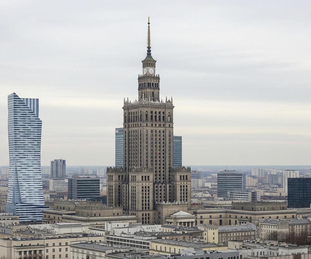 W centrum kadru wznosi się beżowy, monumentalny wieżowiec z iglicą i zegarami na szczycie. Po jego lewej stronie znajduje się nowoczesny, wysoki budynek o geometrycznych, niebiesko-białych fasadach, a po prawej widać kolejny, smukły wieżowiec z anteną na dachu. Otacza je gęsta zabudowa miejska, złożona z niższych budynków o zróżnicowanej architekturze, rozciągająca się aż po horyzont pod bladym, zachmurzonym niebem.