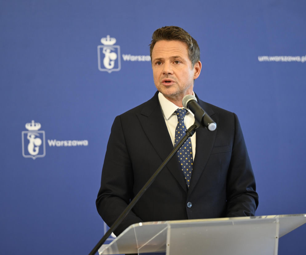 Rafał Trzaskowski przemawiający za mównicą, w tle widoczne logo Warszawy na niebieskiej ścianie, co stanowi kontekst dla inicjatywy odwołania prezydenta, o której można przeczytać więcej na naszym portalu.